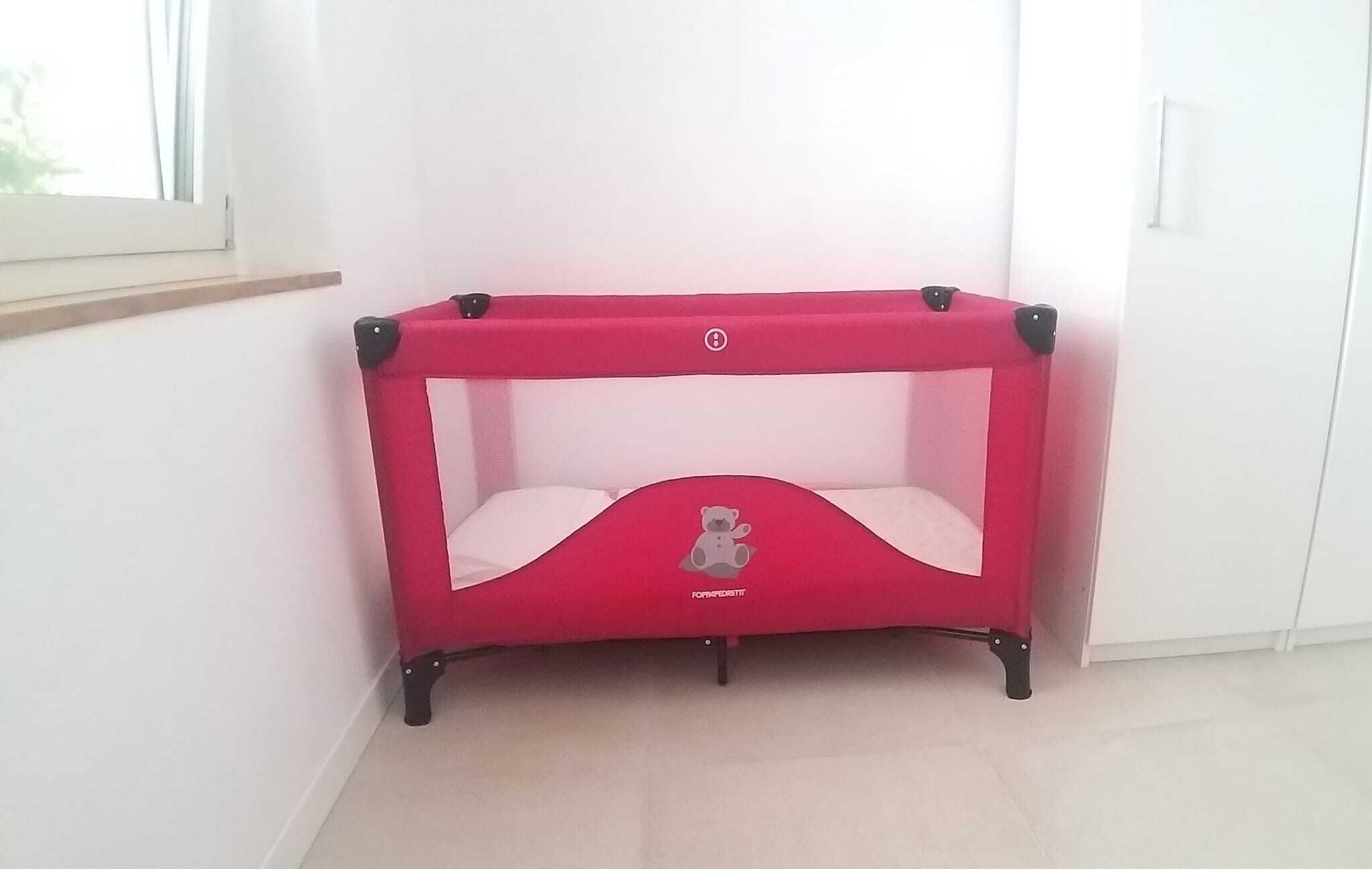 A red baby crib with a bear design, positioned in a corner of a room with white walls and a window.