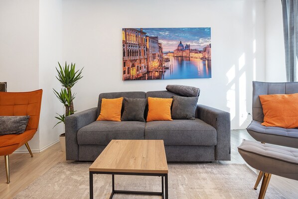 Un salon moderne avec un canapé gris, des coussins orange, une table basse en bois et un tableau représentant un paysage urbain accroché au mur.