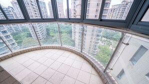 Un balcon incurvé doté d’un sol carrelé, entouré de rampes de verre, qui offre une vue sur une zone urbaine dense, avec de hauts immeubles et des espaces verts.