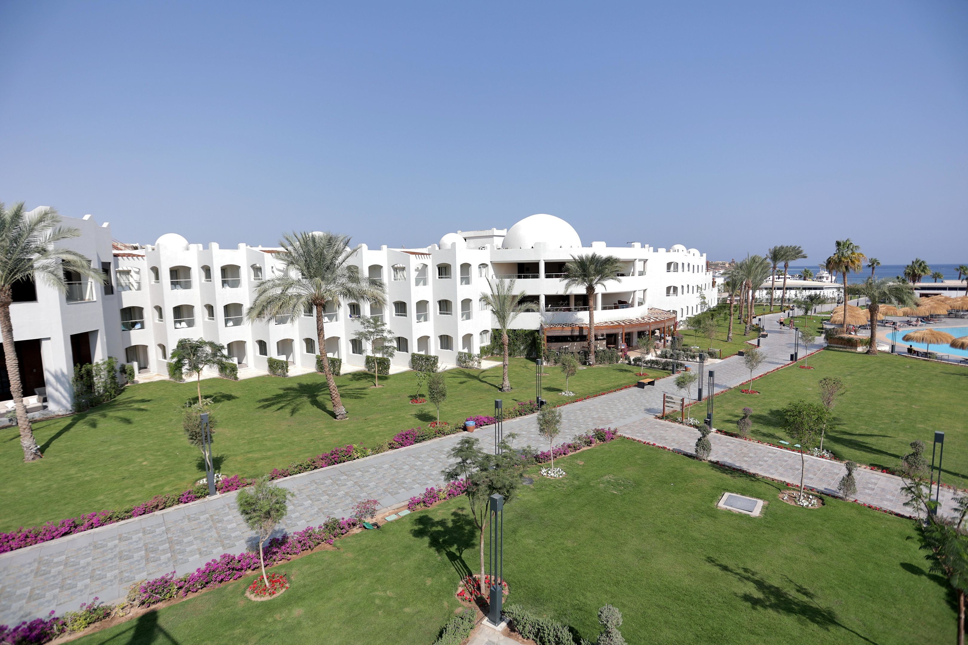 A large white hotel with multiple floors and a central dome, surrounded by well-maintained lawns and palm trees.