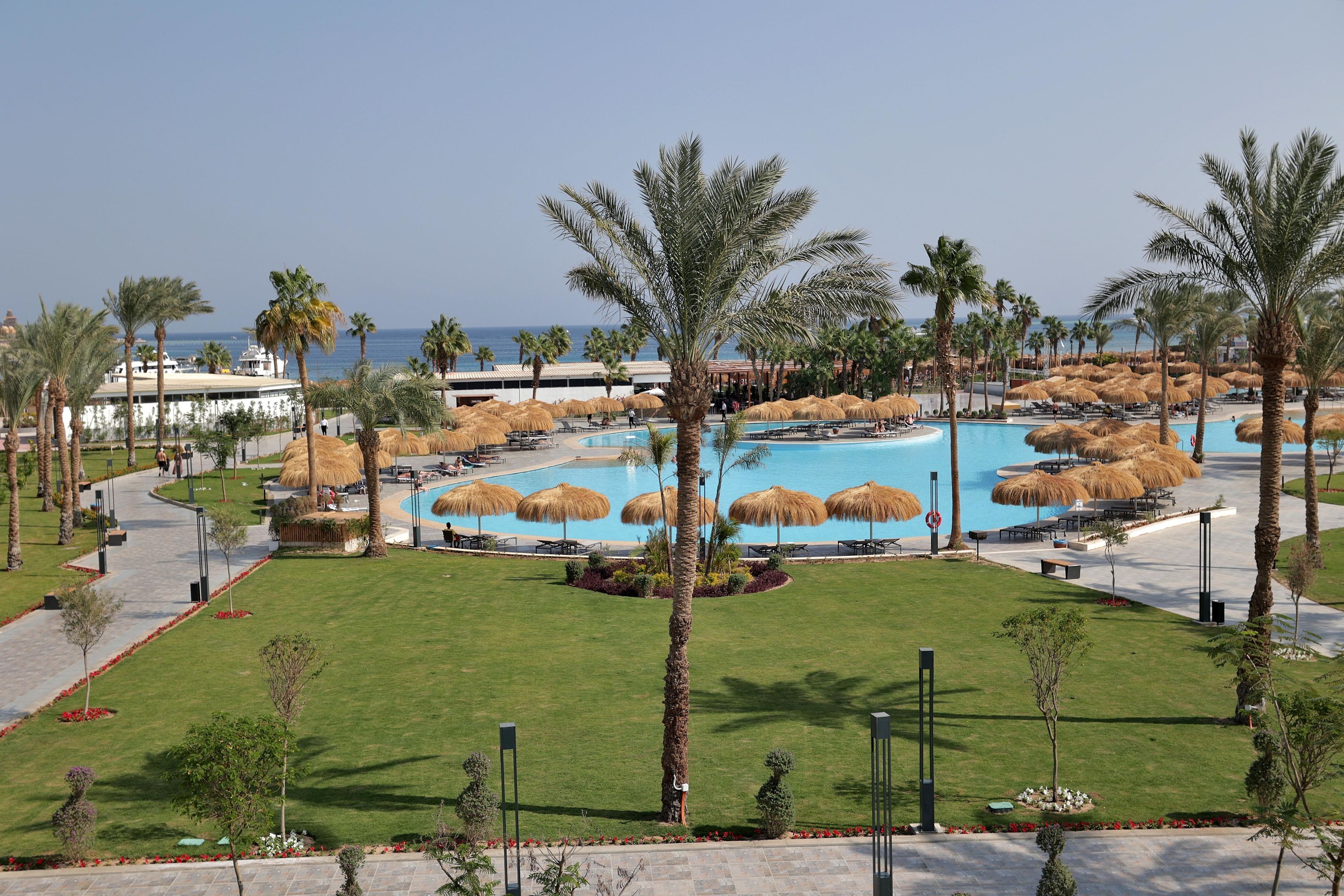 A resort with a central pool area surrounded by palm trees and thatched umbrellas, a clear view of the sea, and a well-maintained lawn.