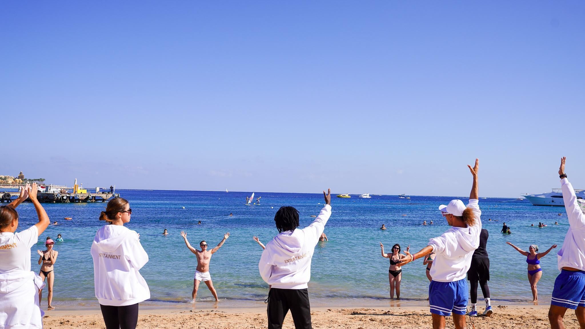 Una playa con gente disfrutando del agua, barcos a lo lejos y un cielo despejado.