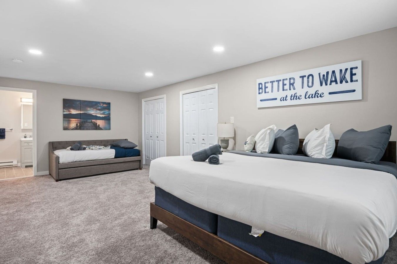 A bedroom with a large bed, a bedside table, a nightstand, a wall-mounted artwork, and a door leading to a bathroom.