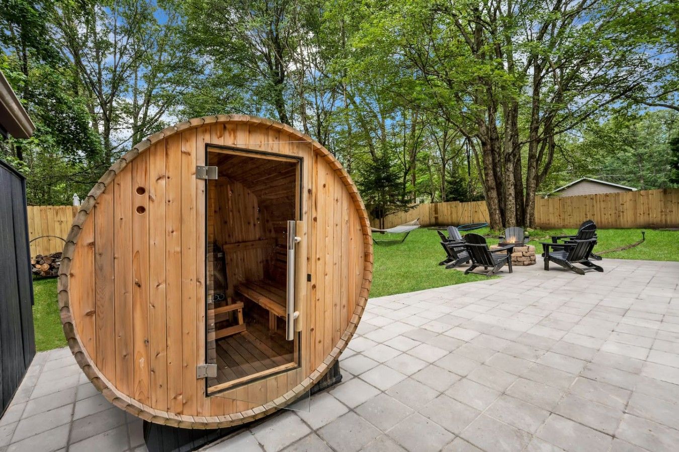 A wooden sauna structure with a door open, a patio area with chairs and a fire pit, and a wooden fence in the background.