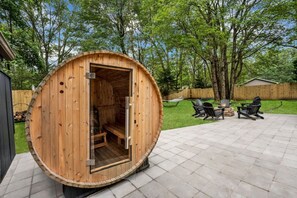 A wooden sauna structure with a door open, a patio area with chairs and a fire pit, and a wooden fence in the background.