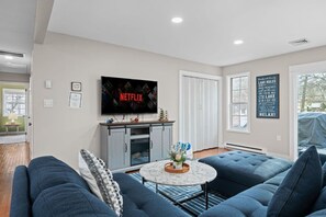 A modern living room with a large flat-screen TV mounted on the wall, a grey cabinet below it, a blue sofa set, a marble coffee table, and a window with a view of trees. - Lake House: Hottub, Sauna, Firepit, Games! (Albrightsville)