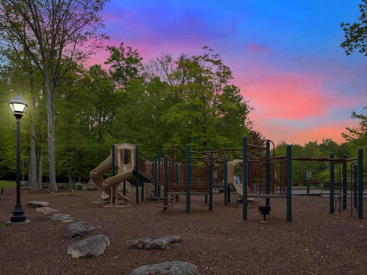 A playground with climbing structures and a slide, surrounded by trees and a street lamp.