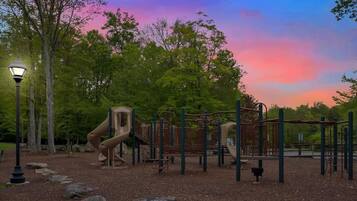 A playground with climbing structures and a slide, surrounded by trees and a street lamp.
