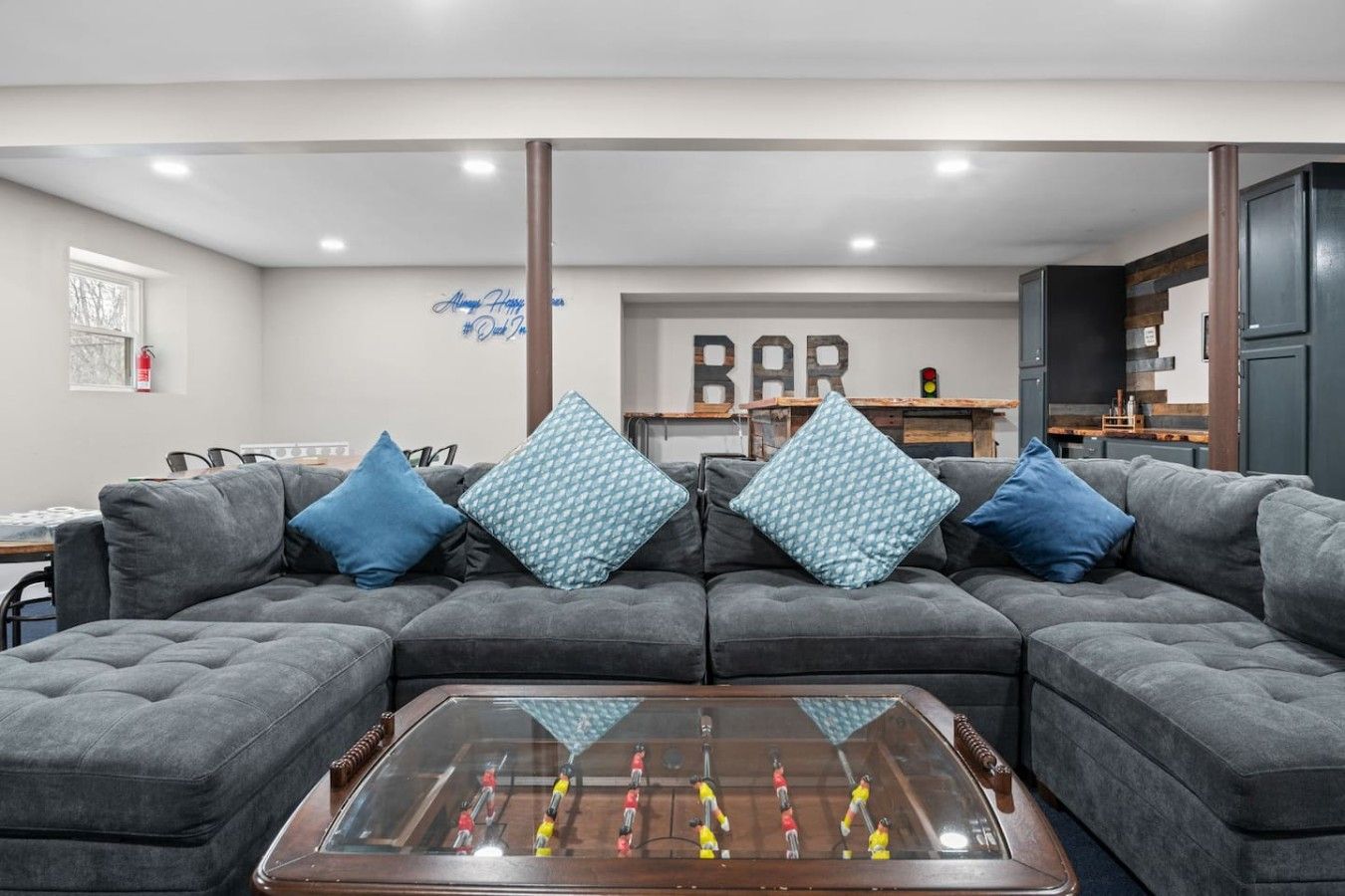 A living room with a sectional sofa, a foosball table, and a bar area in the background.