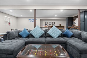 A living room with a sectional sofa, a foosball table, and a bar area in the background. - Lake House: Hottub, Sauna, Firepit, Games! (Albrightsville)