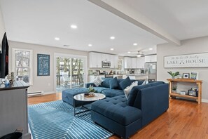 A modern living room with a sectional sofa, a coffee table, and a view of the kitchen. - Lake House: Hottub, Sauna, Firepit, Games! (Albrightsville)