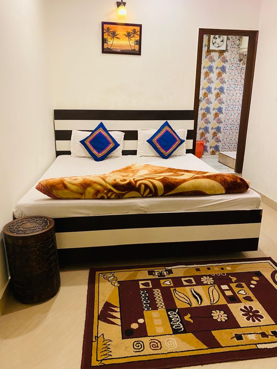 Una cama con un edredón estampado, dos almohadas con un diseño azul y rosa, un taburete de madera y una alfombra estampada en el suelo.
