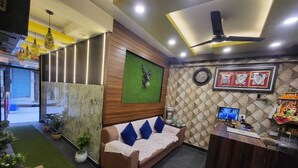 A reception area with a wooden reception desk, a sofa with blue cushions, a green wall with a clock, and a ceiling fan. - Hotel New Pratap (Haridwar)