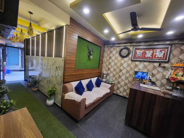 Un espace d’accueil comprenant un comptoir de réception en bois, un canapé avec des coussins bleus, un mur vert orné d’une horloge et un ventilateur de plafond.