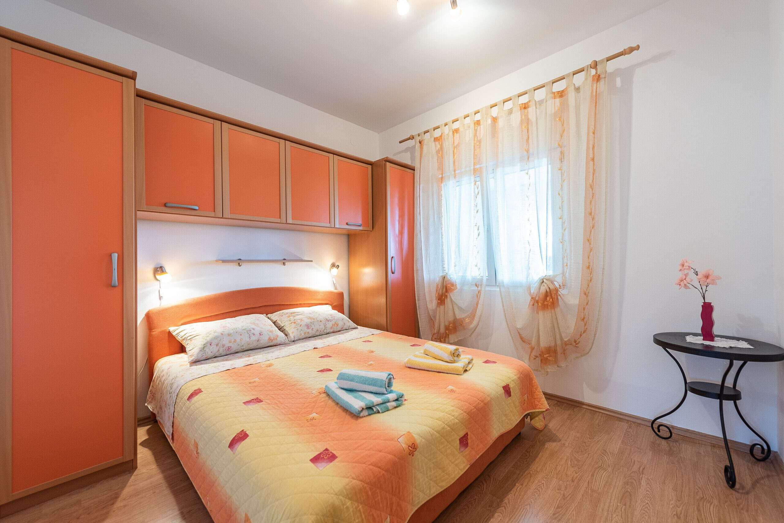 A bedroom with a bed, orange cabinets, a bedside table with a vase, and a window with curtains.