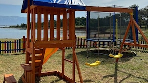 A wooden play structure with a blue roof and swings, located in a grassy area with a lake and mountains in the background. - Beachside Holiday Home Marija (Lozice)