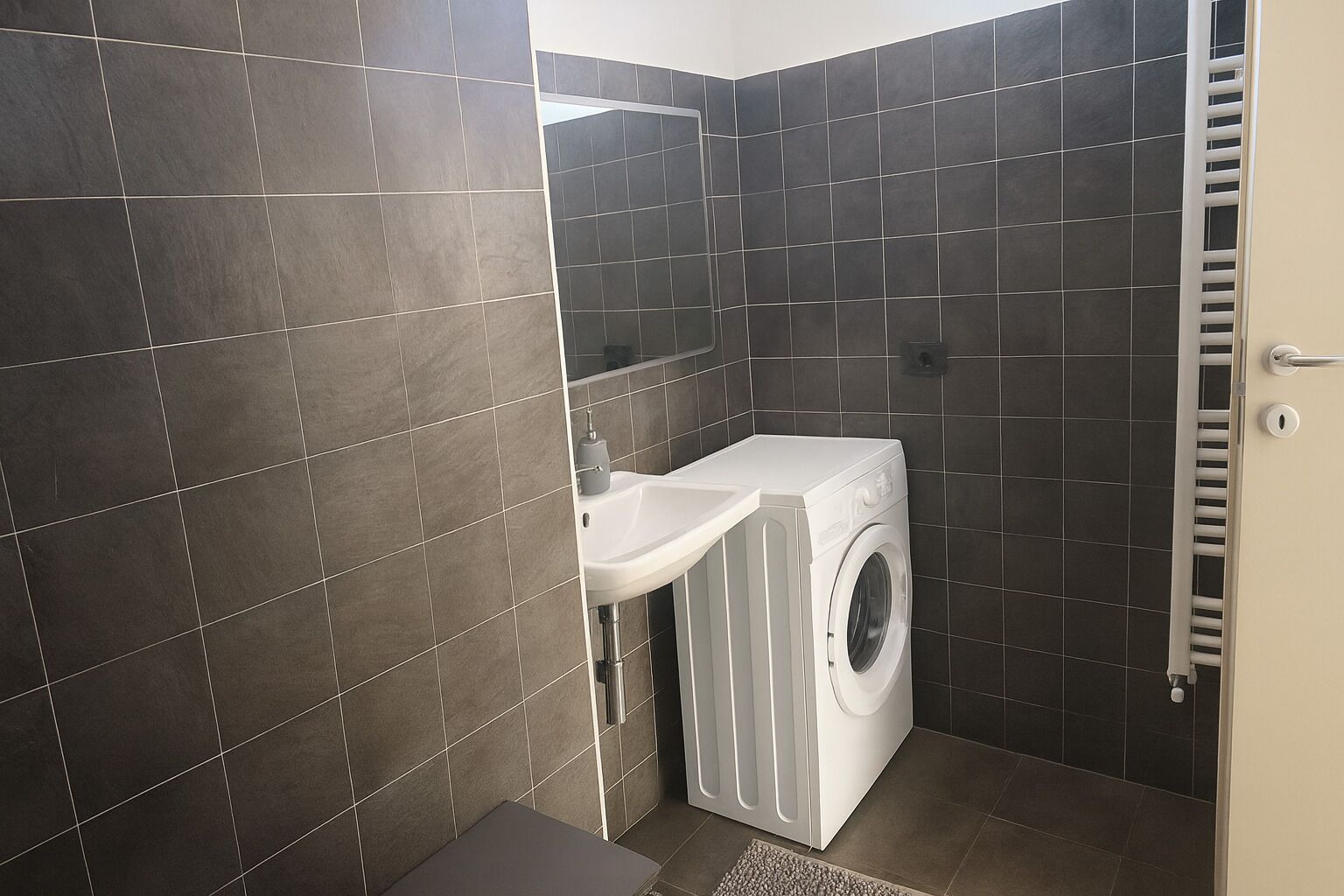 A compact bathroom with dark tiles, a white sink, and a front-loading washing machine.