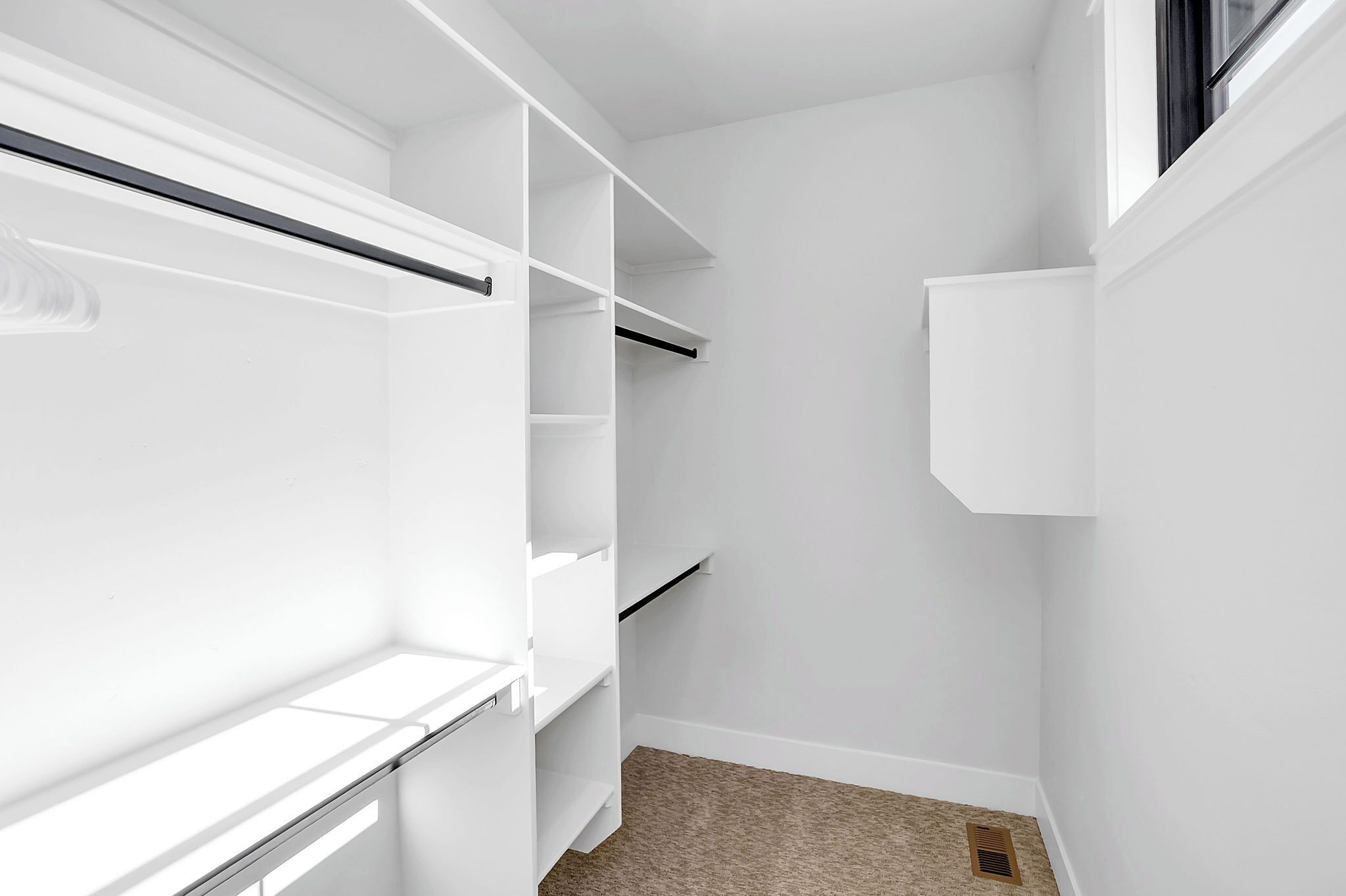 Un armario blanco con estantes, espacio para colgar ropa, una ventana pequeña y suelo alfombrado.