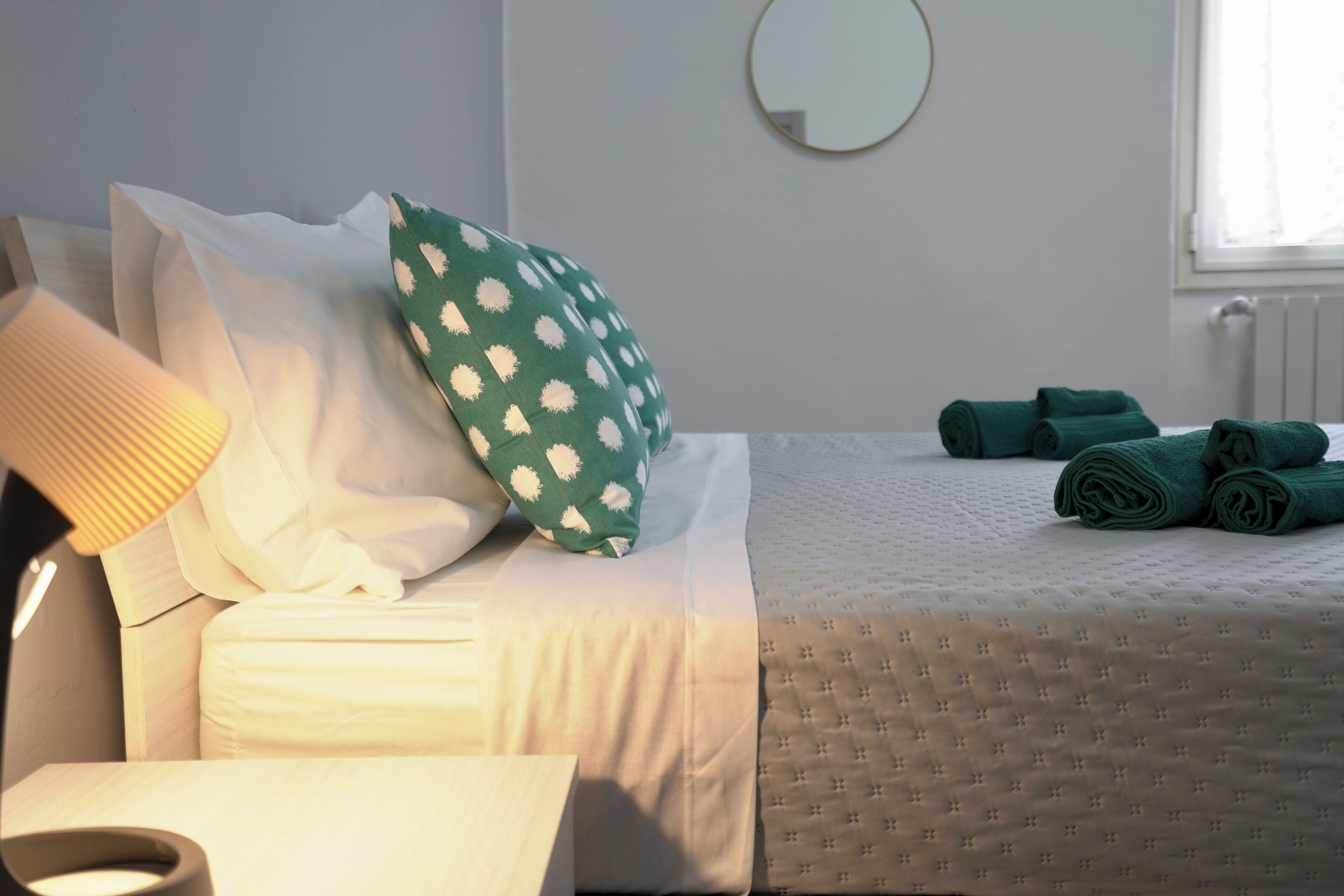 A neatly made bed with white and green pillows, a rolled-up towel, and a green towel on the floor.