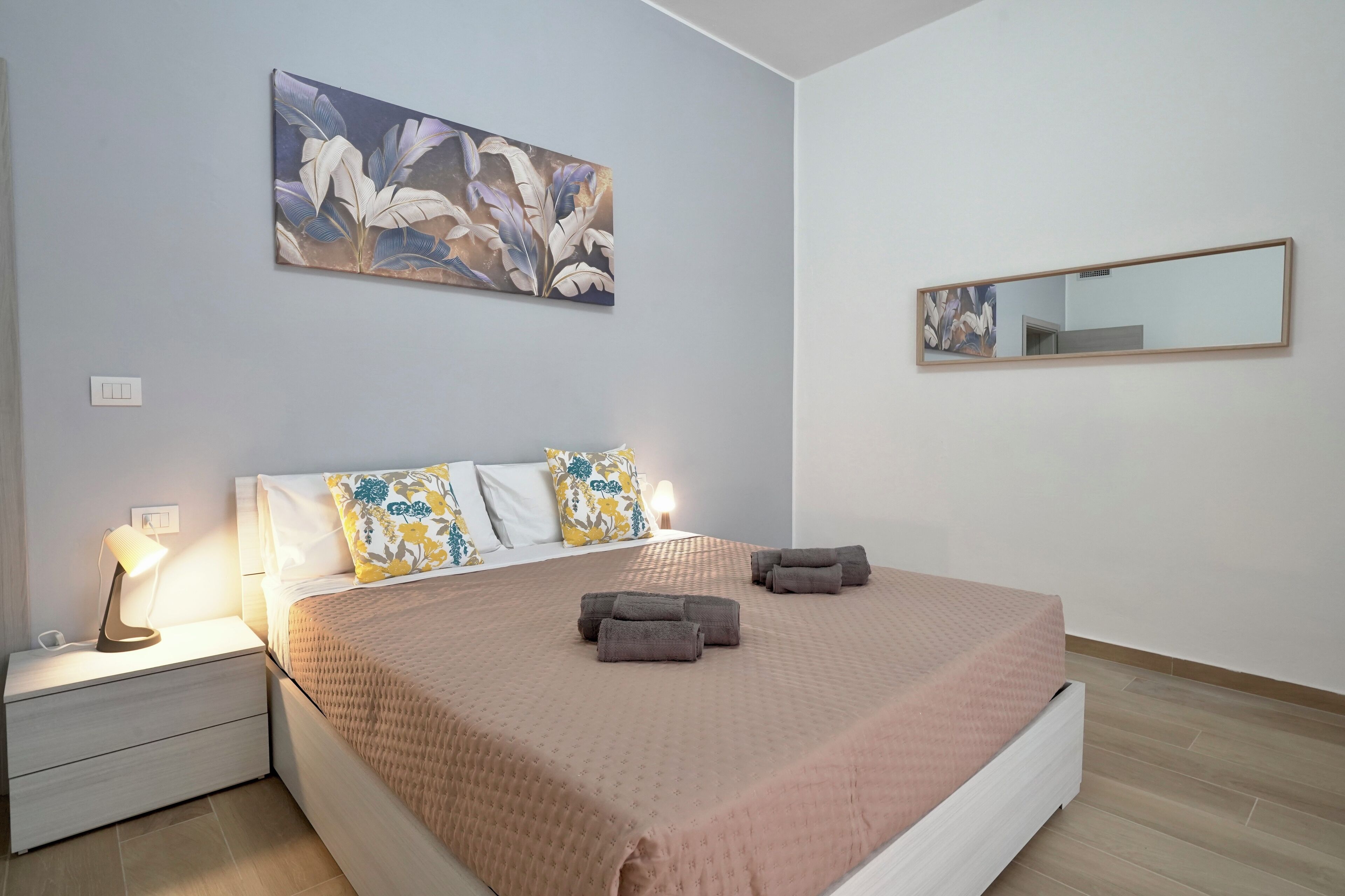 Een net opgemaakt bed met een roze dekbed, een nachtkastje met een lamp en een spiegel aan de muur.