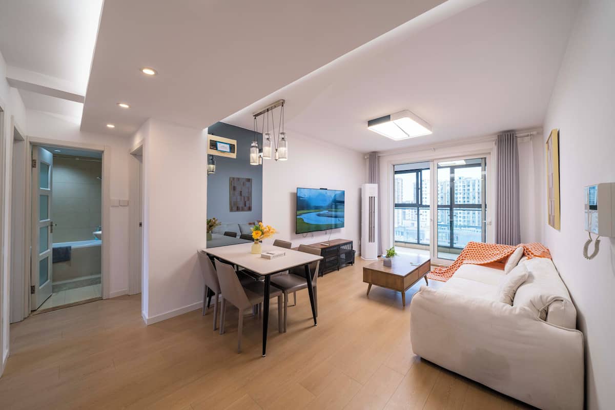 A modern living room with a sofa, a dining table with chairs, a TV, and a large window with a view.