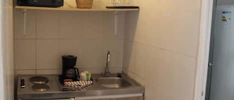 Een compacte kitchenette met een magnetron, een klein spoelgedeelte, een koffiezetapparaat, en een rode ovenwanten die aan de kast hangt.