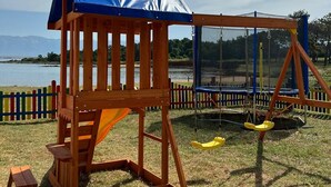 A wooden play structure with a blue roof and swings, located in a grassy area with a lake and mountains in the background. - Beachside Holiday Home Manuela (Lozice)
