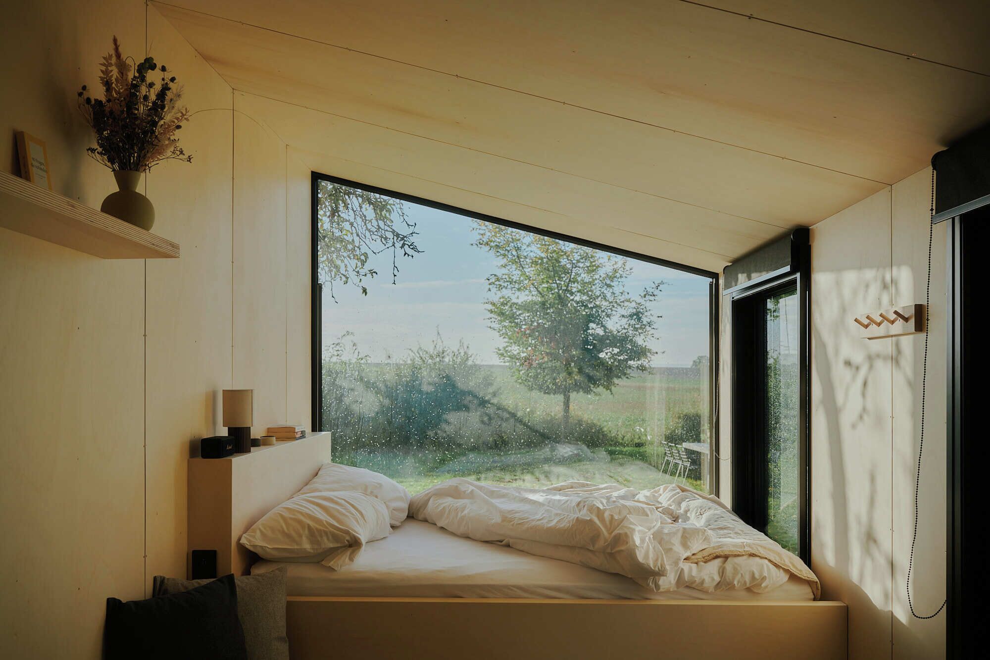 A bedroom with a large window, a bed, a bedside table, and a wall-mounted shelf with a vase.