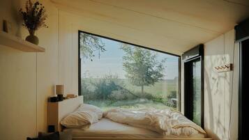 A bedroom with a large window, a bed, a bedside table, and a wall-mounted shelf with a vase.