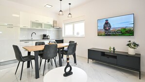 Eine moderne Küche mit weißen Schränken, einem Holztisch und schwarzen Stühlen. Ein Flachbildfernseher an der Wand zeigt ein landschaftliches Außenbild.