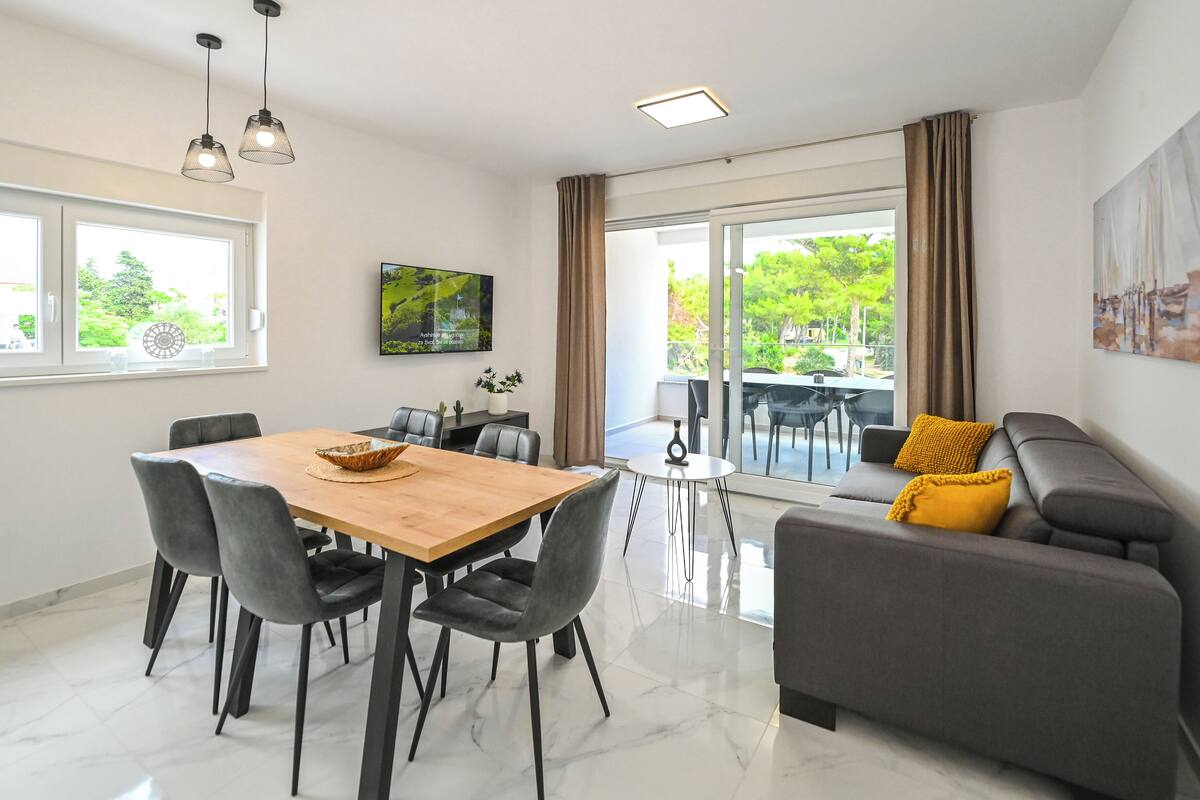 A modern living room with a dining area, a grey sofa with yellow cushions, and a large window offering a view of greenery.
