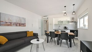 Ein modernes Wohnzimmer mit Sofa, Couchtisch und Essbereich mit Stühlen und Tisch. Im Hintergrund befindet sich eine Küche mit Schränken und einem Fenster.