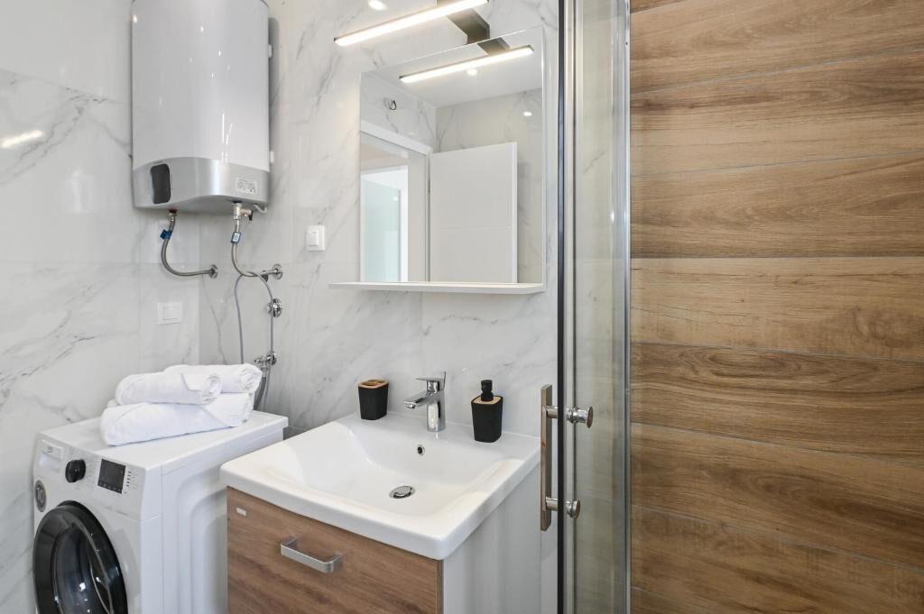 A modern bathroom with a marble wall, a wooden panel wall, a glass shower enclosure, a built-in washing machine, and a sink with a wooden cabinet.