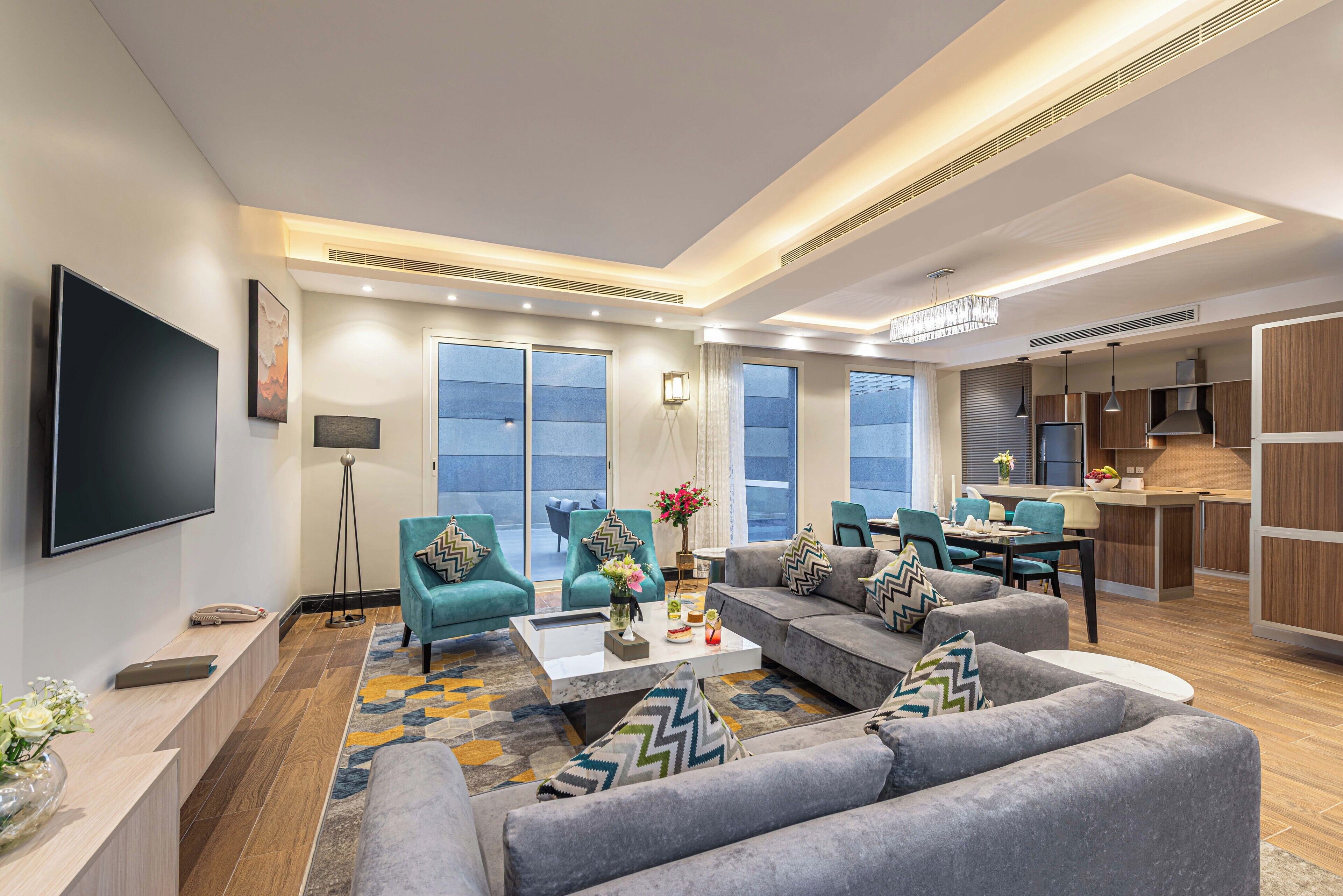 A modern living room with a flat-screen TV, comfortable seating, a coffee table, and a kitchen area.