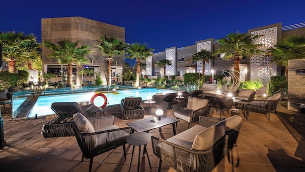 An outdoor pool area with lounge chairs and tables, surrounded by palm trees and modern buildings.