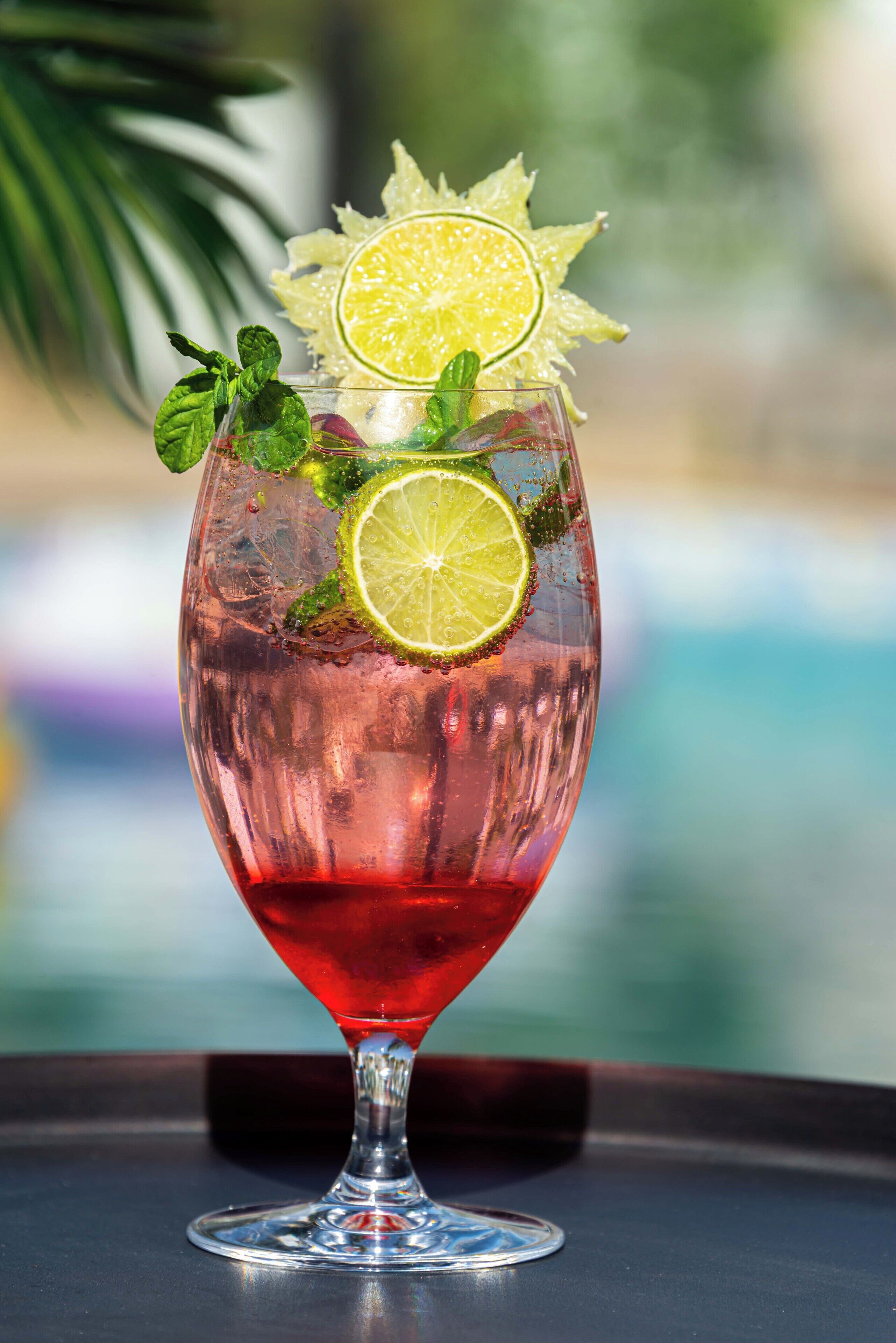 A cocktail glass with a red drink, garnished with lime slices and mint leaves, placed on a table with a blurred background.