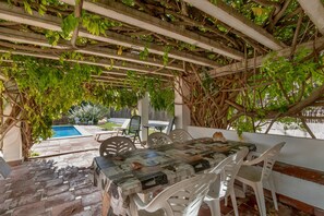 A covered patio with a table and chairs, a pool in the background, and a pergola with climbing plants.