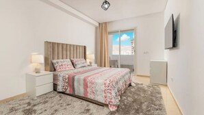 A modern bedroom with a large bed, a nightstand, a TV mounted on the wall, and a view of the city through the window.