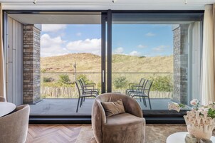 A modern room with a sliding glass door leading to a balcony with chairs and a view of a hilly landscape.