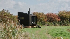 A modern black cabin with a chimney, situated in a grassy field with autumn foliage. - Raus | Cabins mit Feldblick + Saunazugang (Königslutter am Elm)