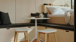 A modern, minimalist room with a bed, a small table with two chairs, and a shelf with books and a vase. - Raus | Cabins mit Feldblick + Saunazugang (Königslutter am Elm)