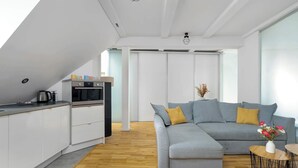 A modern kitchen with white cabinets, a built-in oven, and a coffee maker. There is a grey sectional sofa with a yellow pillow and a small table with a vase of flowers. - Stylish Apartment with Workspace | Close to the Old Zoo (Poznań)