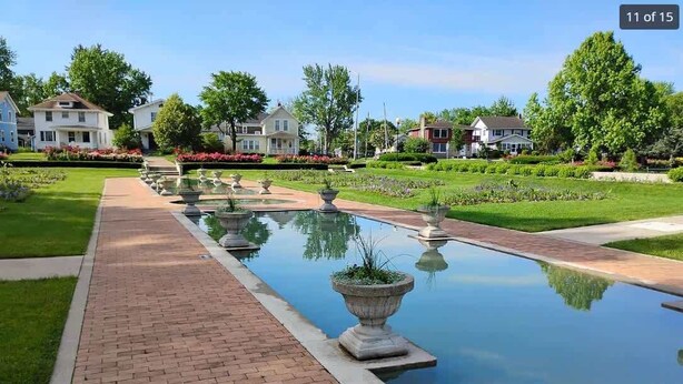 Een rustige tuin met een lang pad van baksteen dat leidt naar een reflecterend watergedeelte, omringd door verzorgde gazons en woonhuizen.