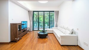 A modern living room with a wooden floor, a white sofa, a TV on a wooden cabinet, and a large window with curtains.
