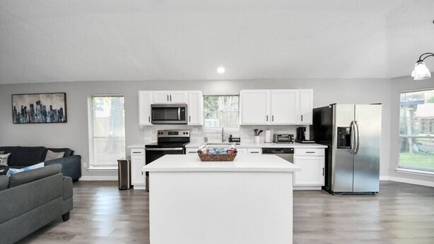 A modern kitchen with white cabinetry, a central island, a refrigerator, and a dining area with a gray sofa.