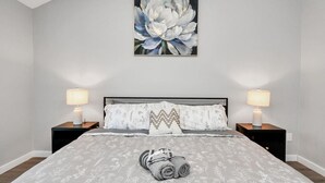 A bedroom with a large bed, two bedside tables with lamps, a wall-mounted painting, and a rolled towel on the bed.