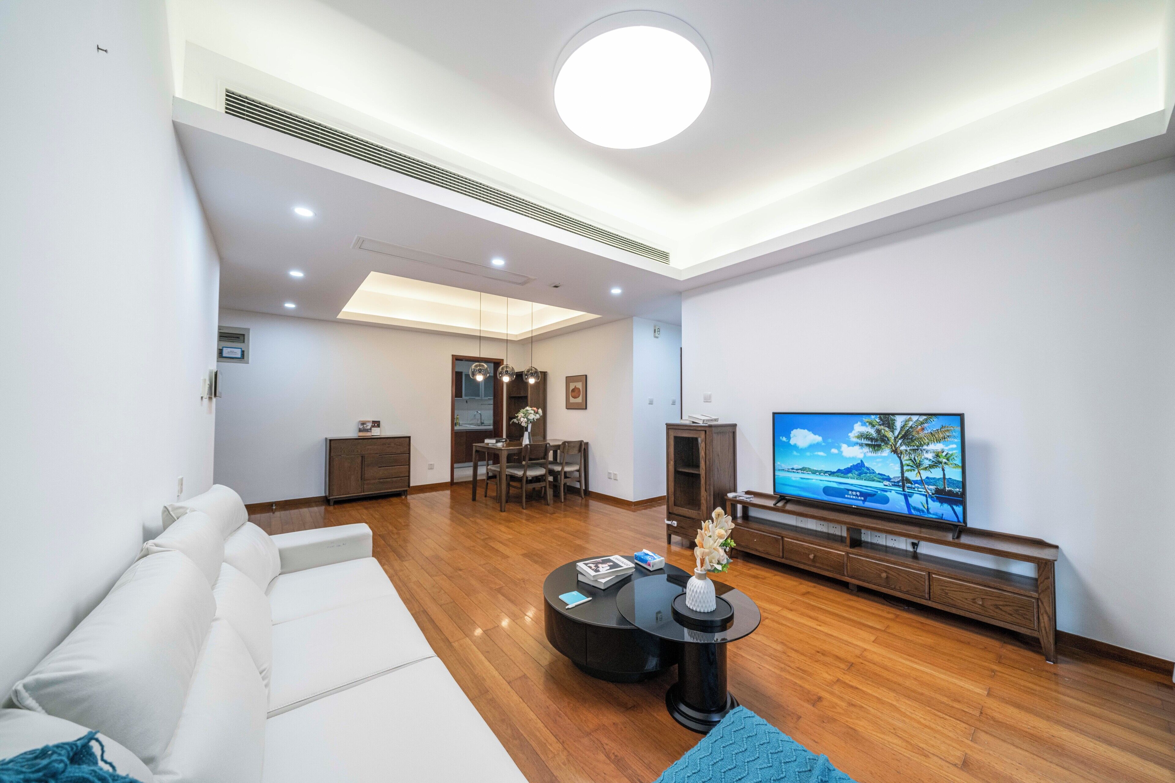 Een moderne woonkamer met een witte bank, een ronde salontafel, een televisie op een houten dressoir en een eethoek met een tafel en stoelen.