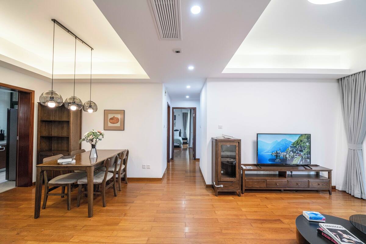 A modern living room with a wooden dining table, chairs, a TV on a wooden stand, and a hallway leading to another room.