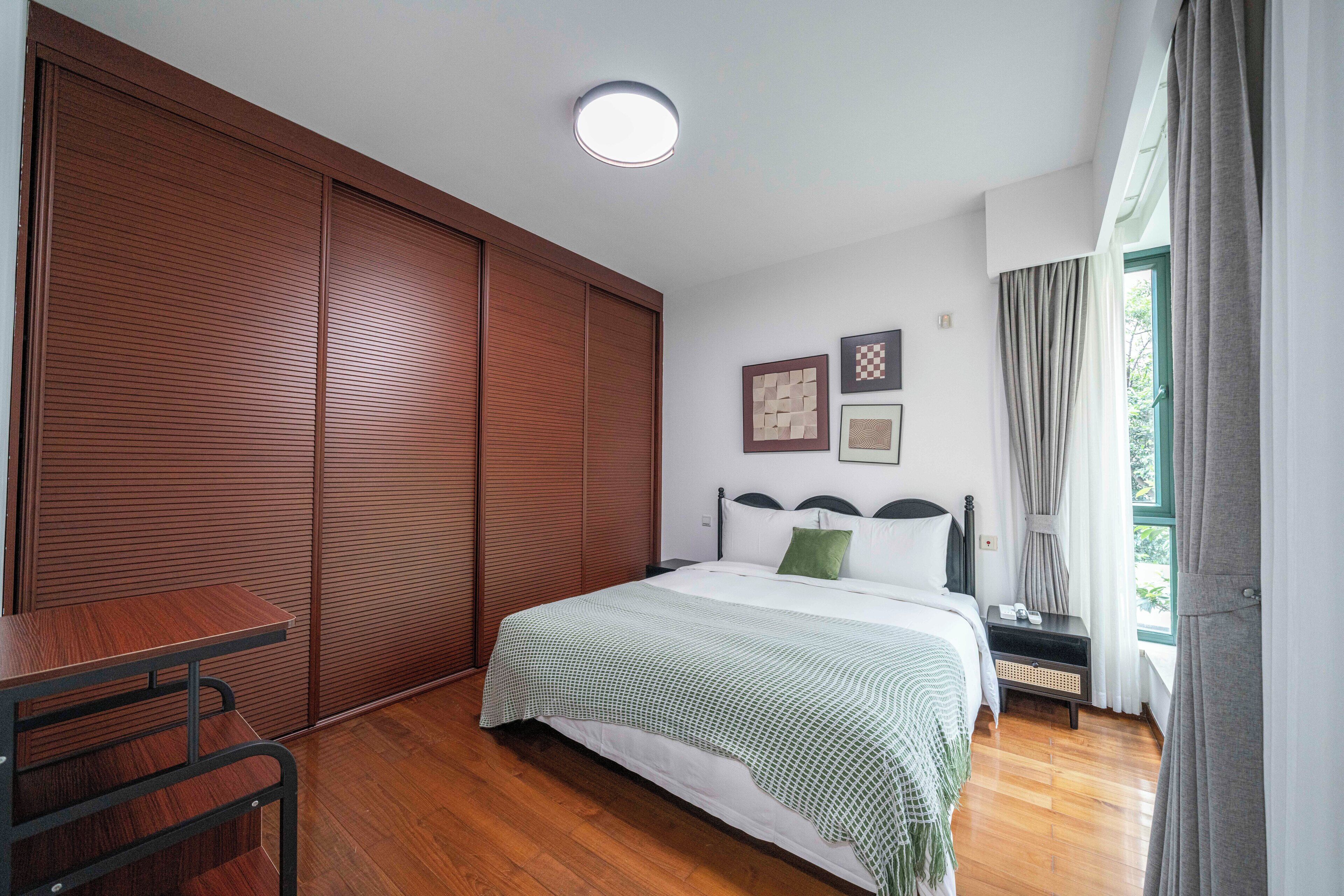 A bedroom with a bed, a wooden wardrobe, a desk, and a window with curtains.