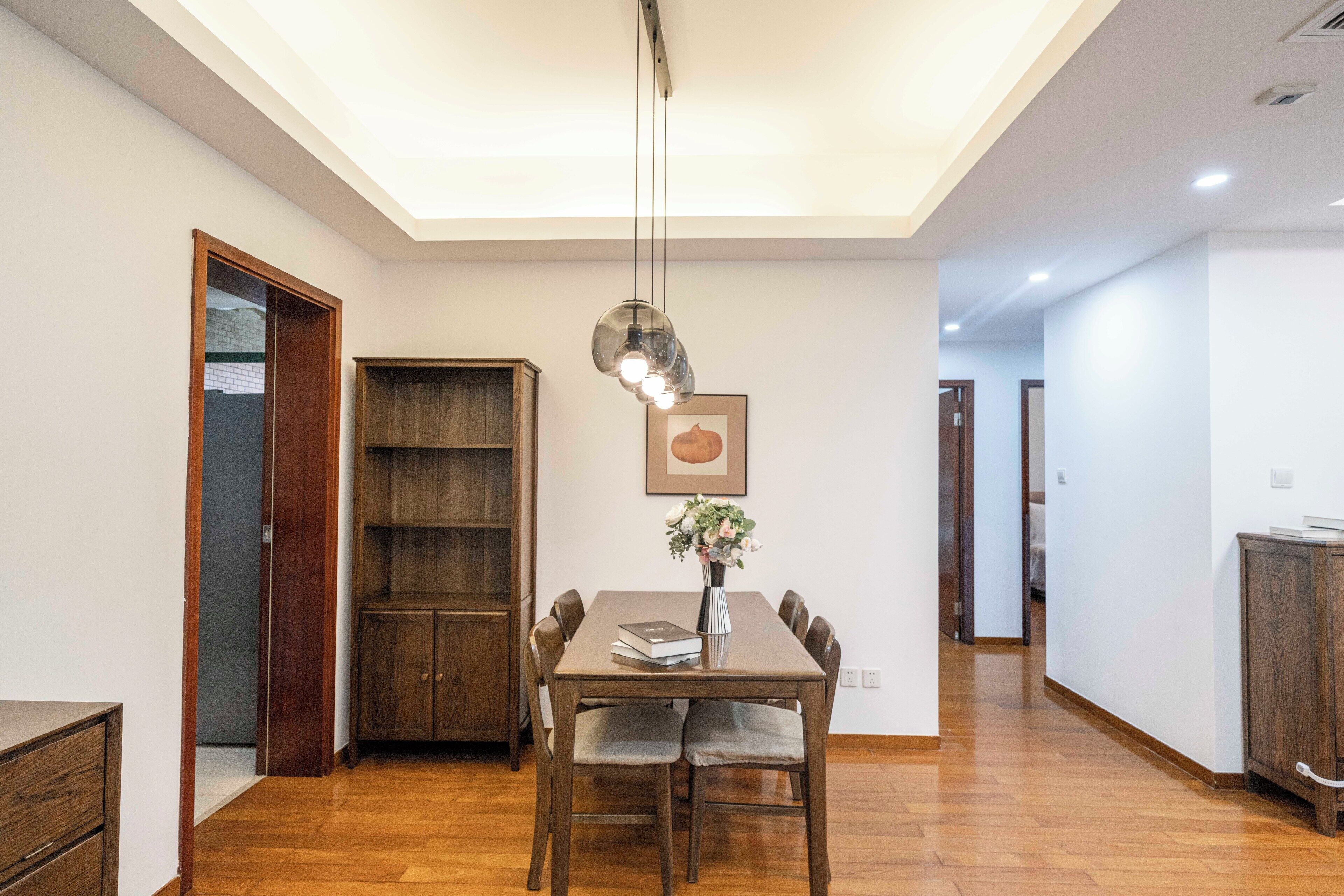 A dining area with a wooden table, chairs, a vase with flowers, and a pendant light.